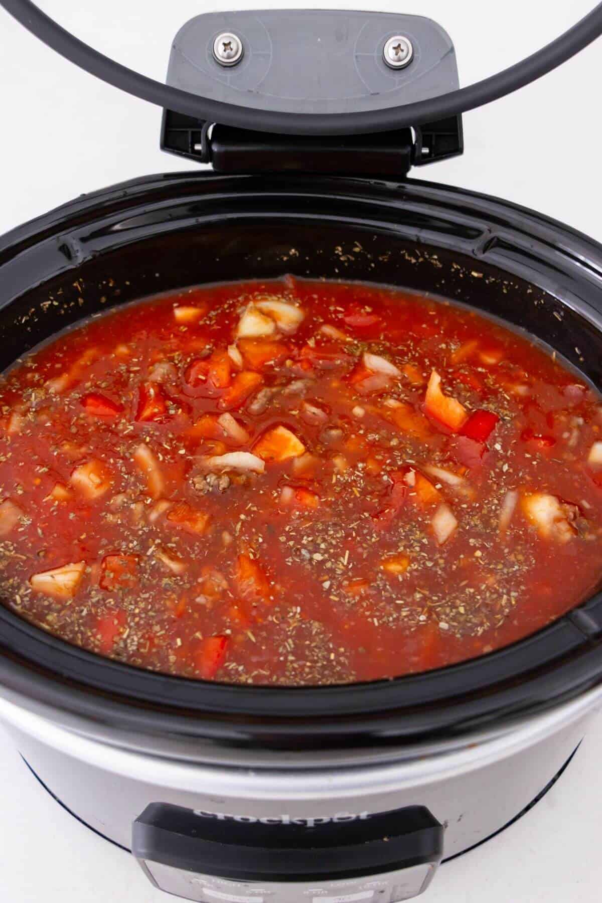 A slow cooker filled with uncooked soup or stew, containing chopped vegetables, ground meat, and tomato-based broth.