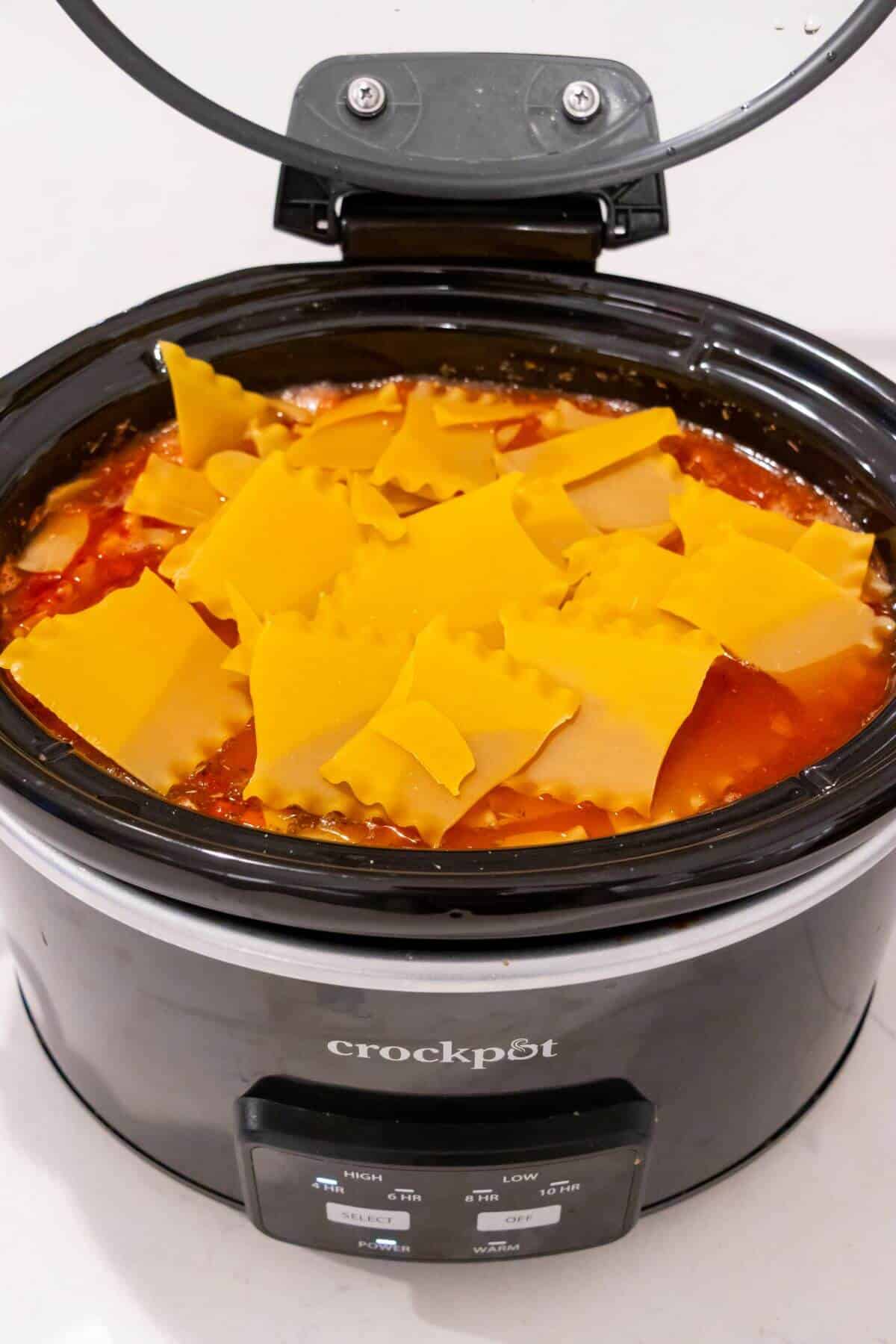 Uncooked lasagna noodles layered over sauce and ingredients in a black Crockpot slow cooker with the lid open.