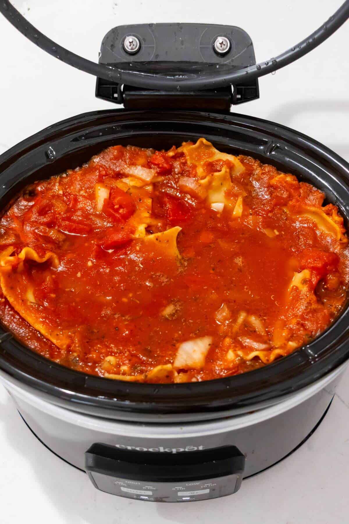 A slow cooker filled with uncooked lasagna noodles, tomato sauce, cheese, and meat, ready to be cooked.