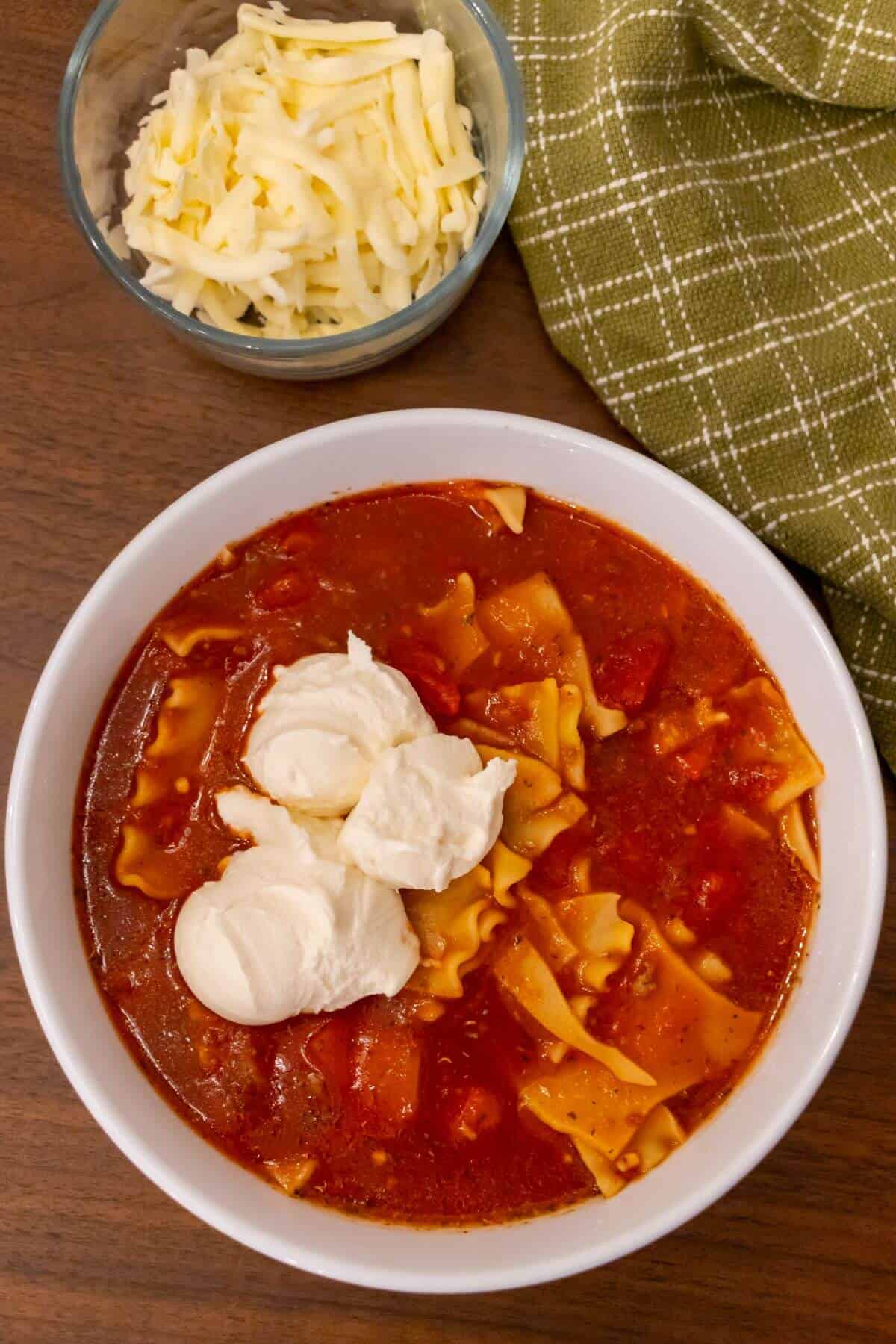 A bowl of lasagna soup topped with three scoops of ricotta cheese, next to a bowl of shredded cheese and a green checkered napkin.