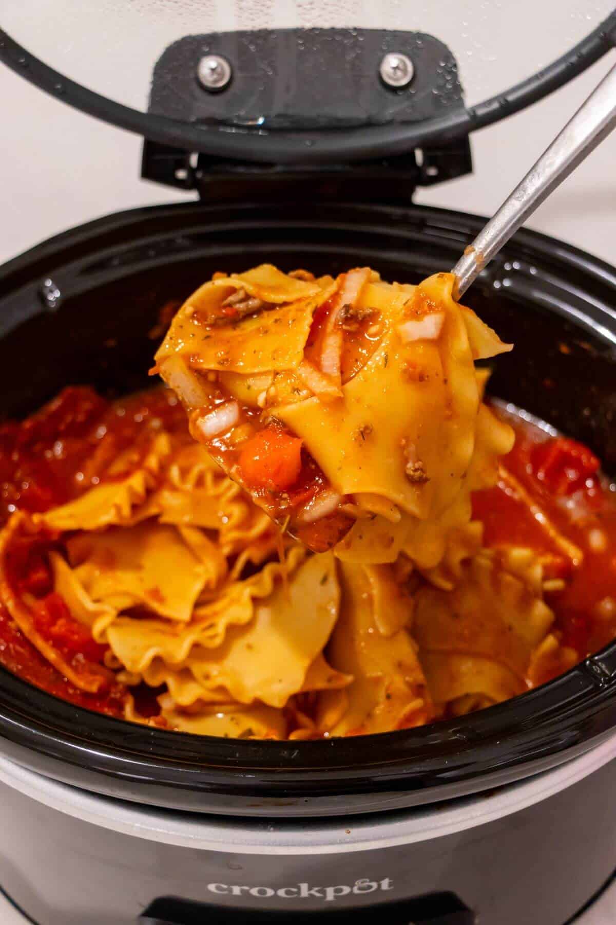 A serving spoon lifts a portion of lasagna from a black Crockpot filled with pasta, tomato sauce, and cheese.