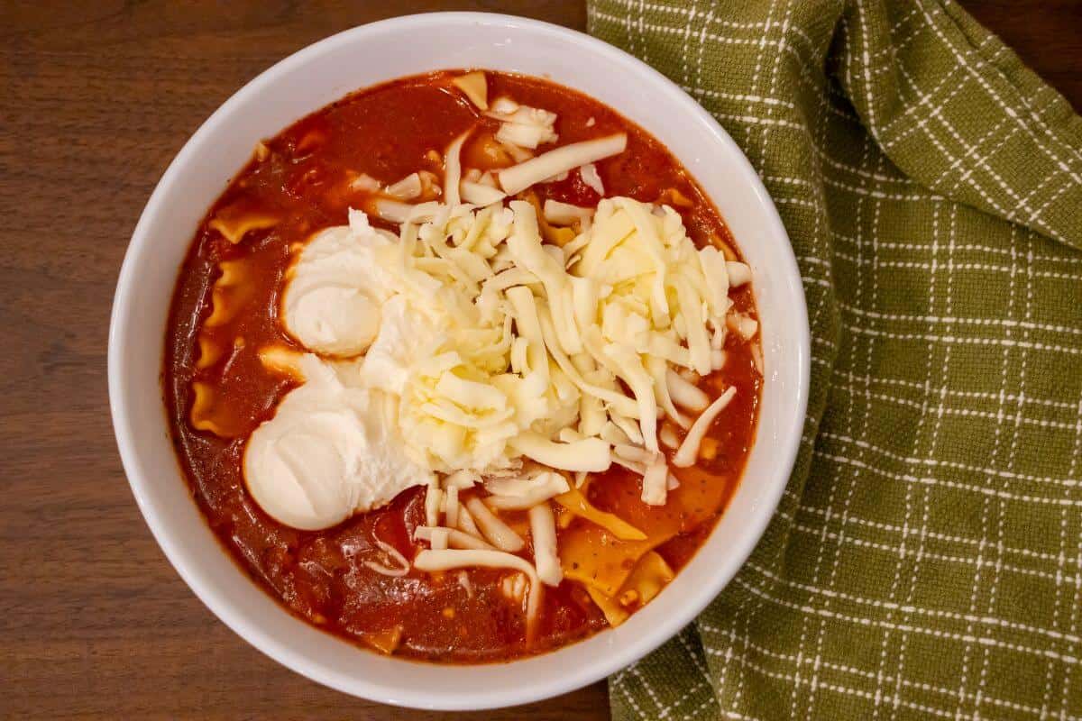 A bowl of tomato-based soup topped with shredded cheese and dollops of sour cream sits on a wooden surface next to a green plaid napkin.