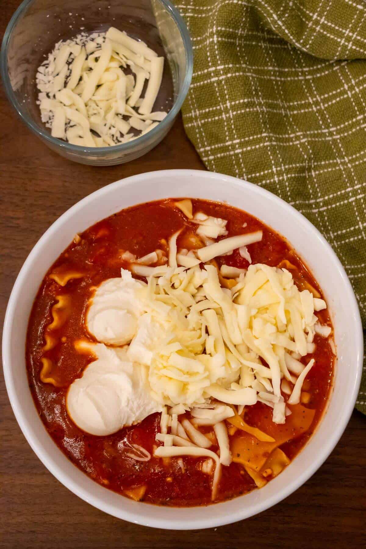 A bowl of lasagna soup topped with shredded cheese and two scoops of ricotta cheese, next to a glass bowl of grated cheese and a green checkered cloth.