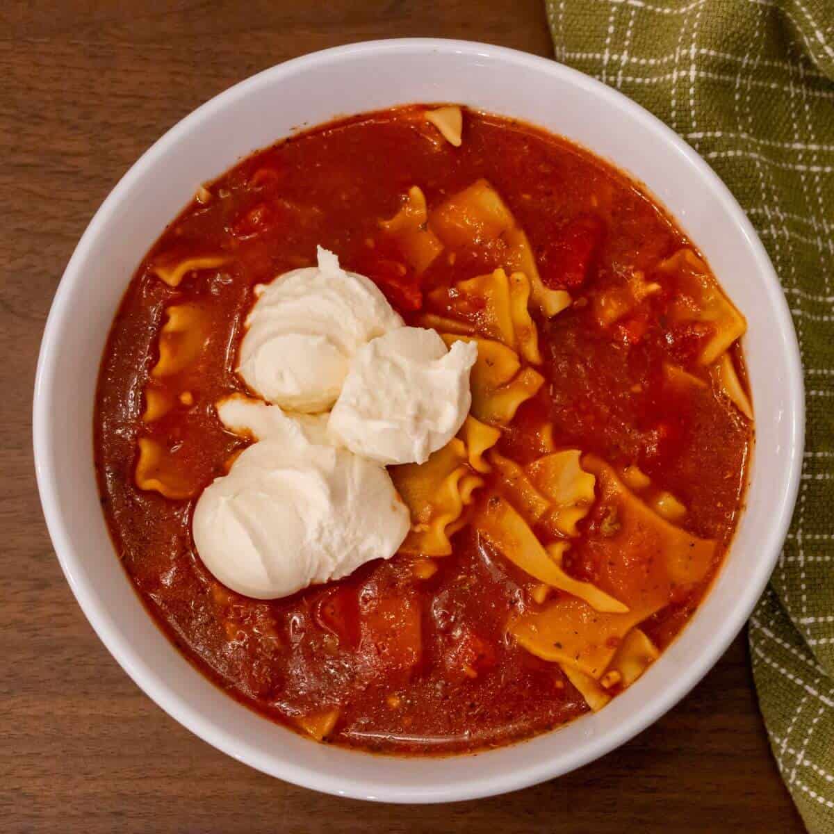 A bowl of tomato-based soup with lasagna noodles and three dollops of ricotta cheese, placed on a wooden table next to a green checkered cloth.