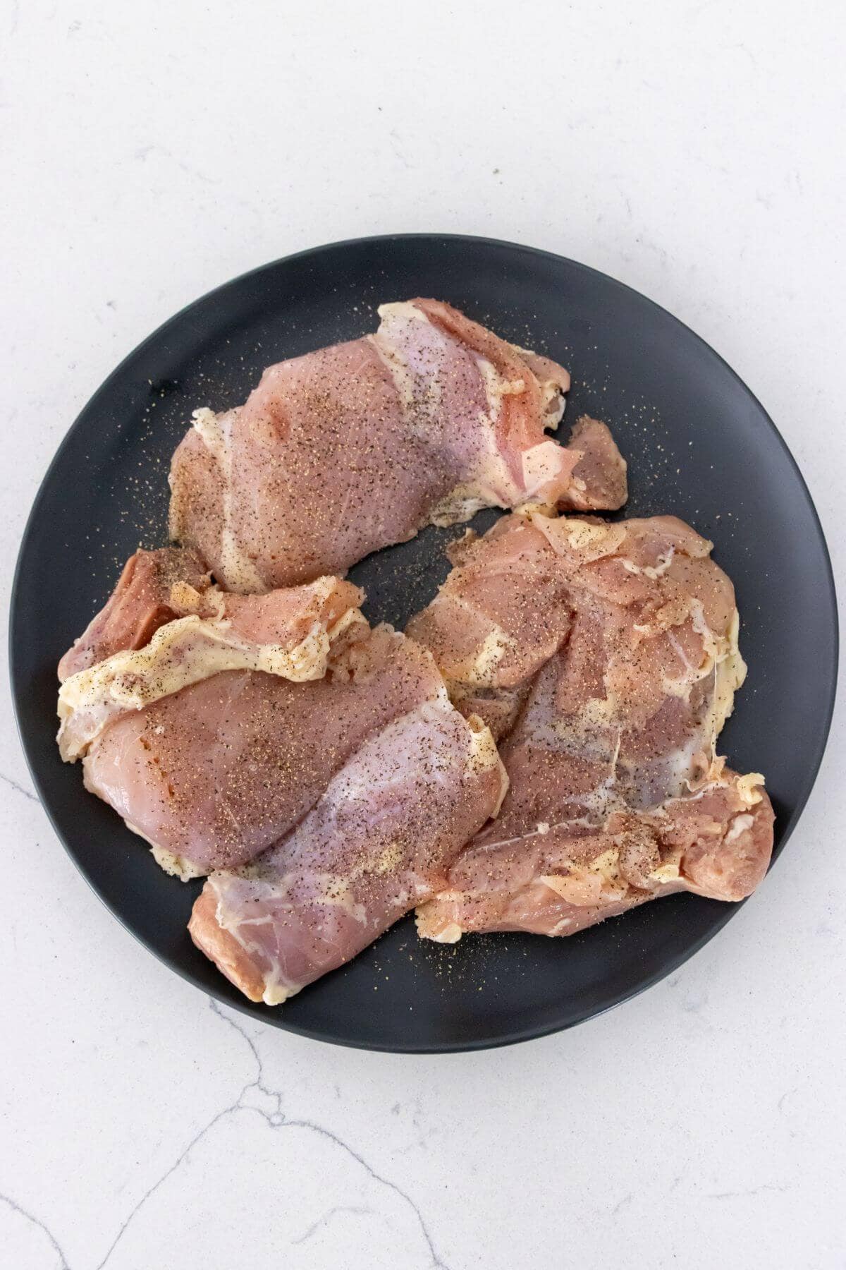 Raw, seasoned chicken thighs arranged on a round black plate, placed on a light-colored countertop.