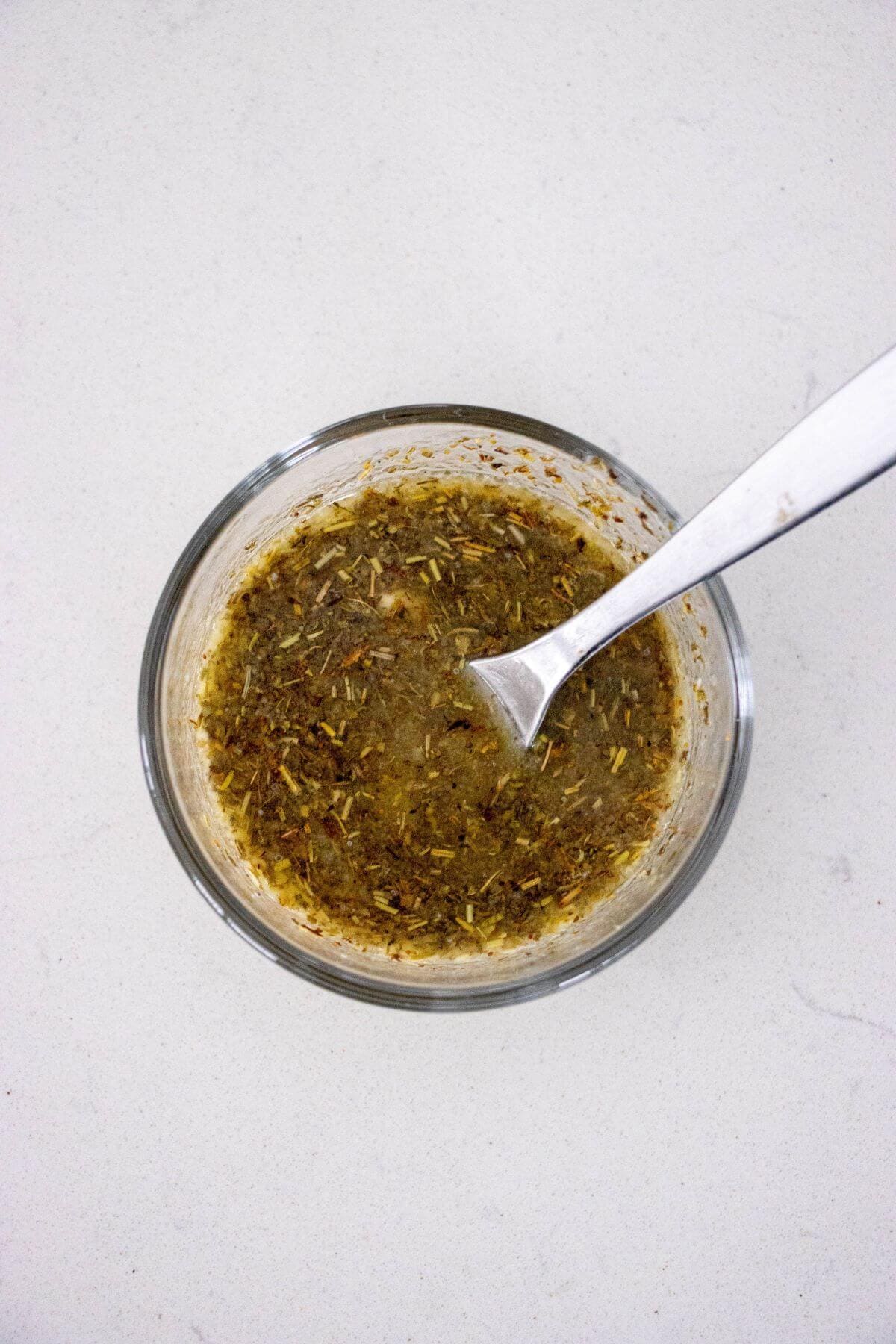 A small glass bowl filled with a mixed spice marinade and a spoon resting inside, placed on a white surface.