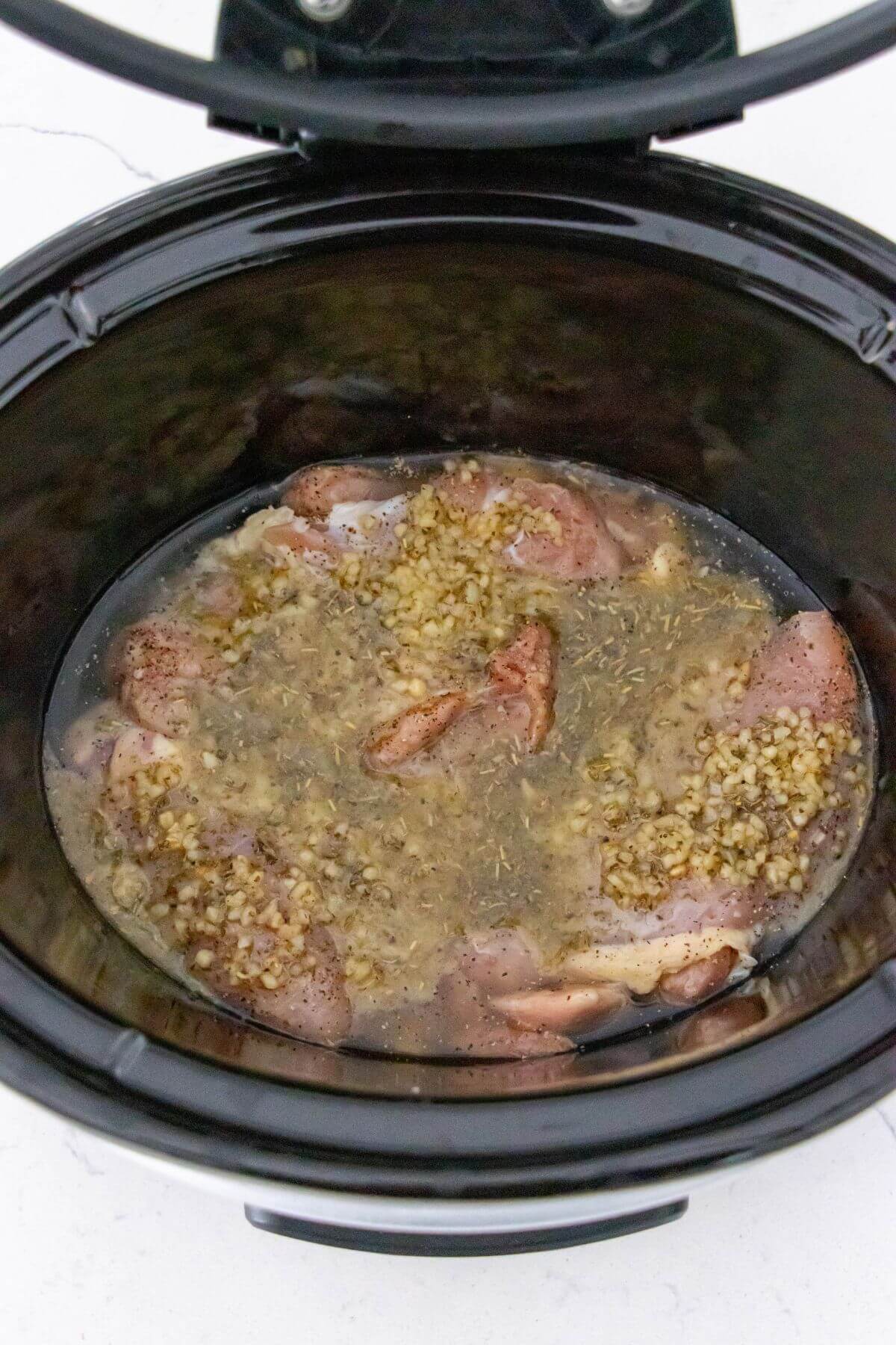 Raw chicken pieces with seasoning and minced garlic in liquid inside a slow cooker, ready to be cooked.