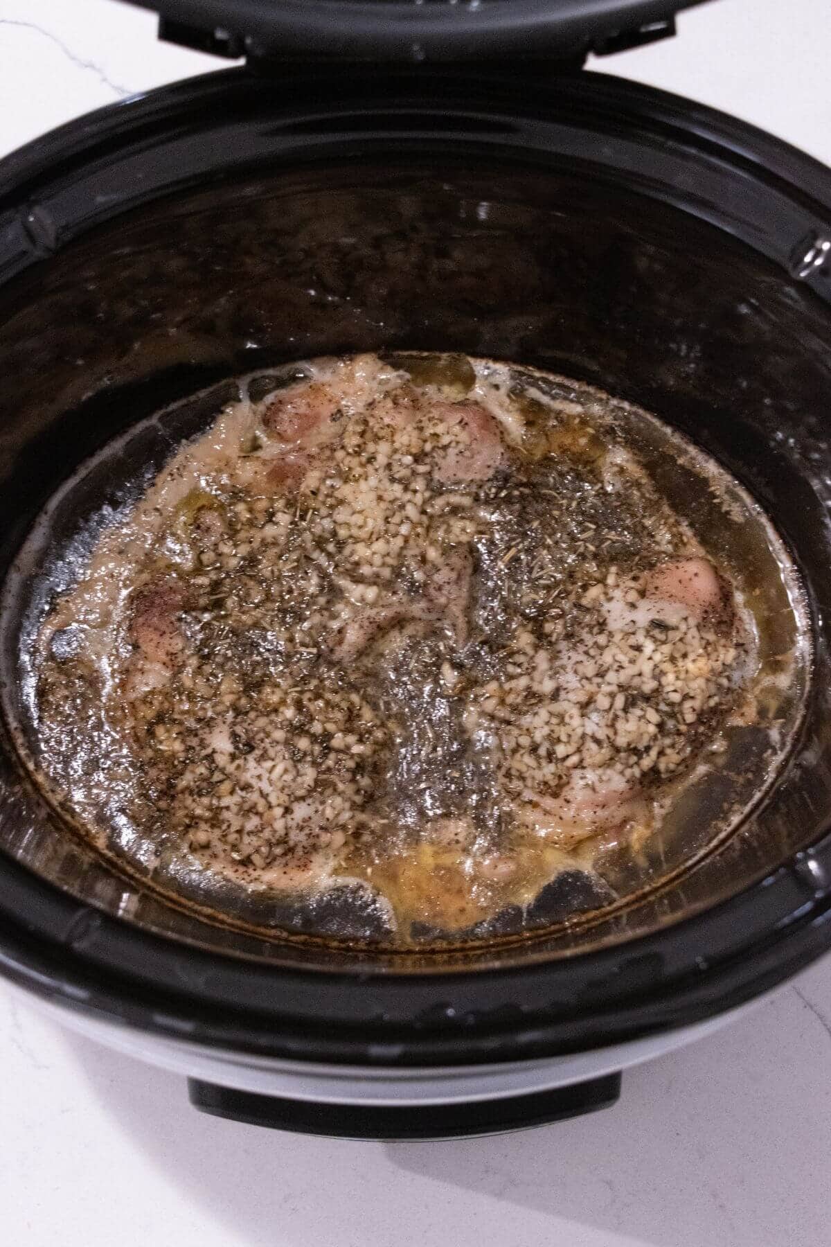 Chicken pieces seasoned with herbs and minced garlic cooking in broth inside a slow cooker.