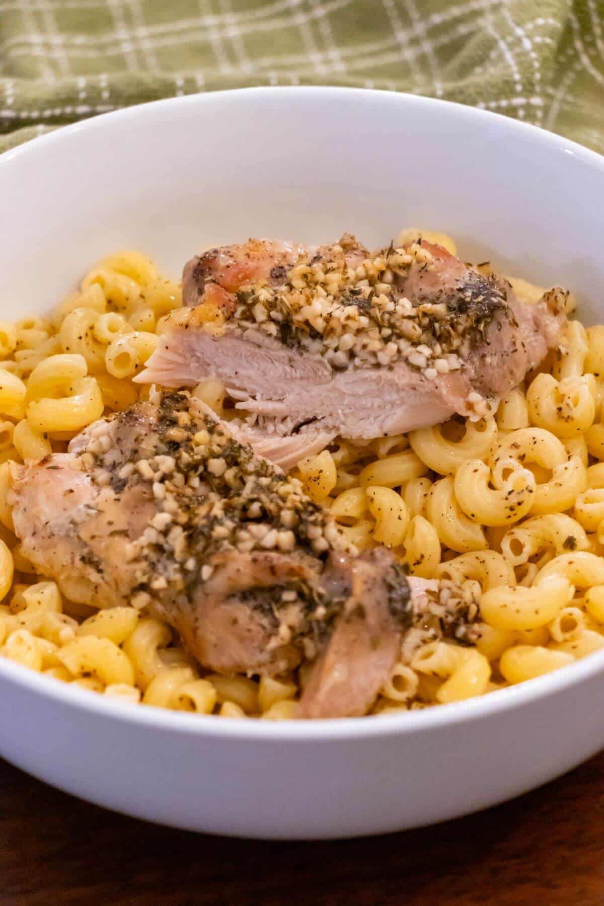 A white bowl filled with macaroni pasta topped with pieces of baked chicken garnished with chopped garlic and herbs.
