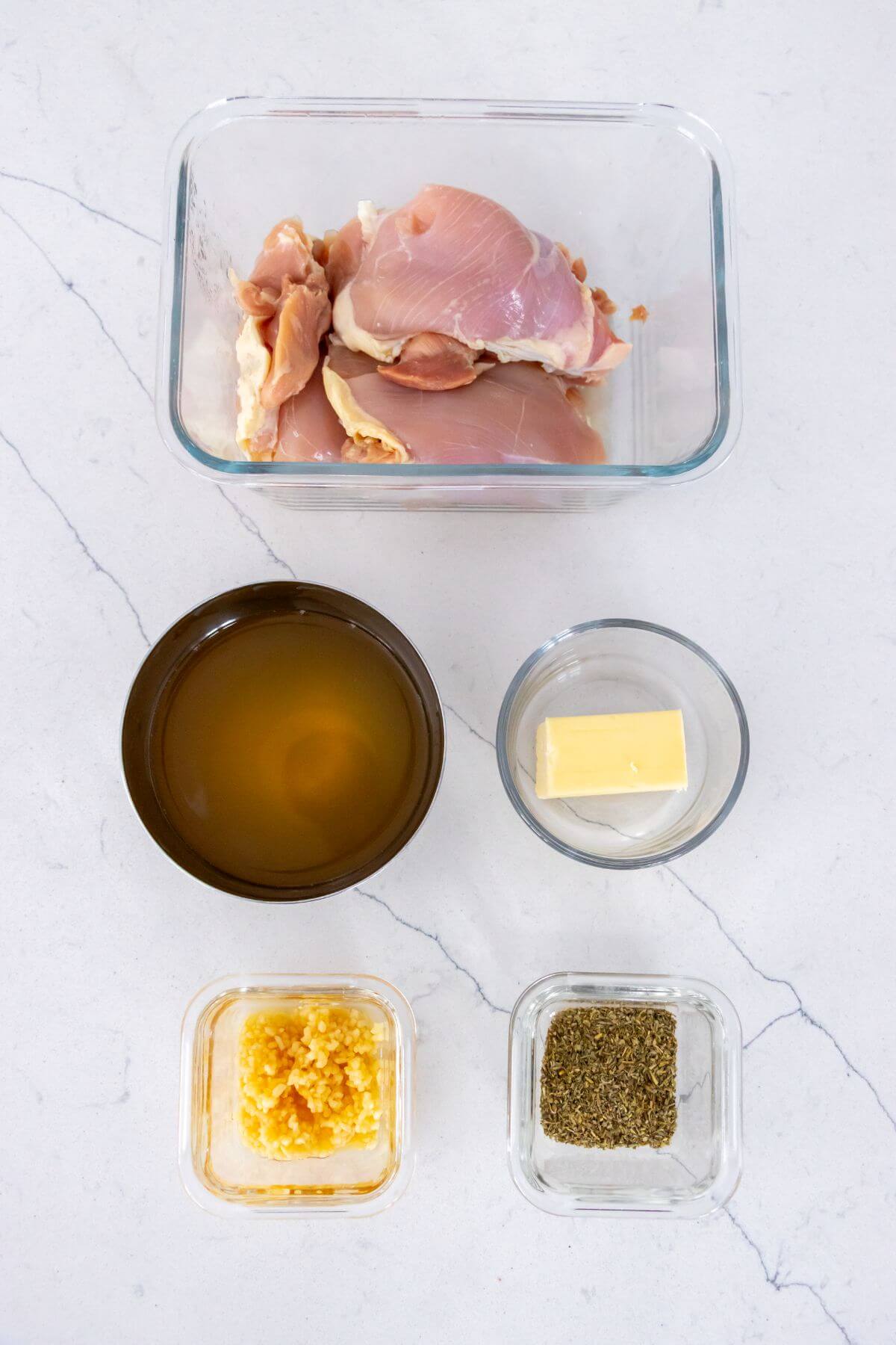 Five ingredients on a white surface: raw chicken thighs, chicken broth, a piece of butter, minced garlic, and Italian seasoning in separate bowls.