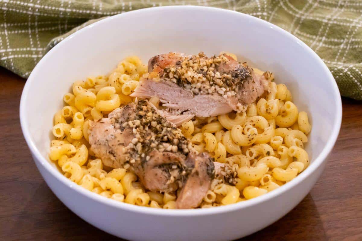 A white bowl filled with macaroni pasta topped with two pieces of seasoned and cooked chicken, placed on a wooden table with a green cloth in the background.