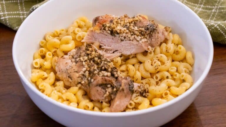 A white bowl filled with elbow macaroni pasta topped with two pieces of seasoned, baked chicken on a wooden table.