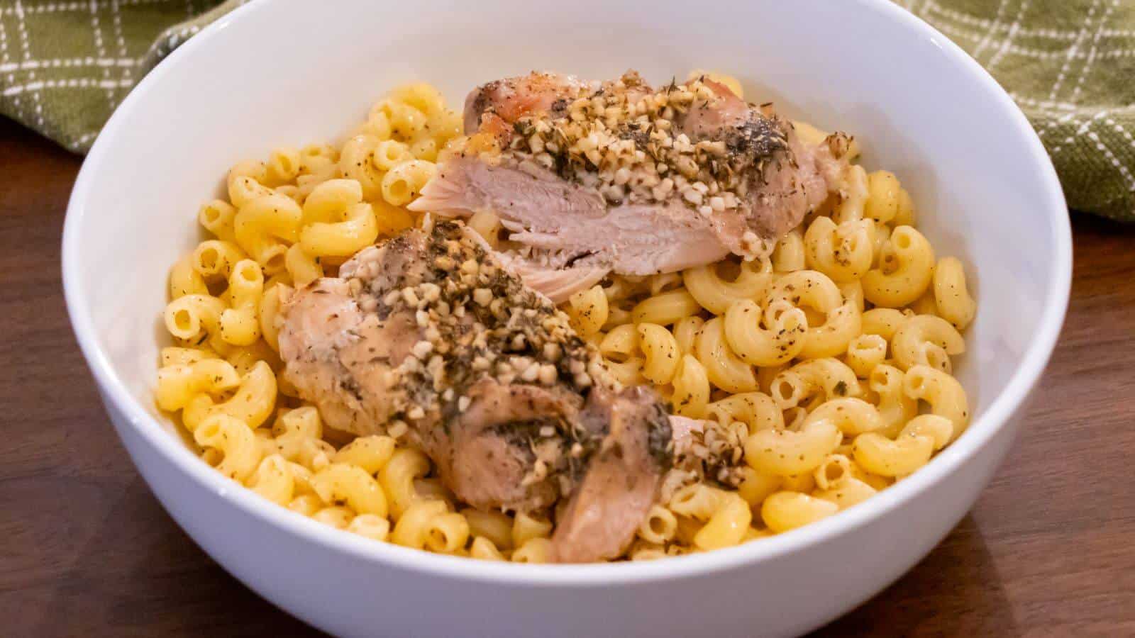 A white bowl filled with elbow macaroni pasta topped with two pieces of seasoned, baked chicken on a wooden table.