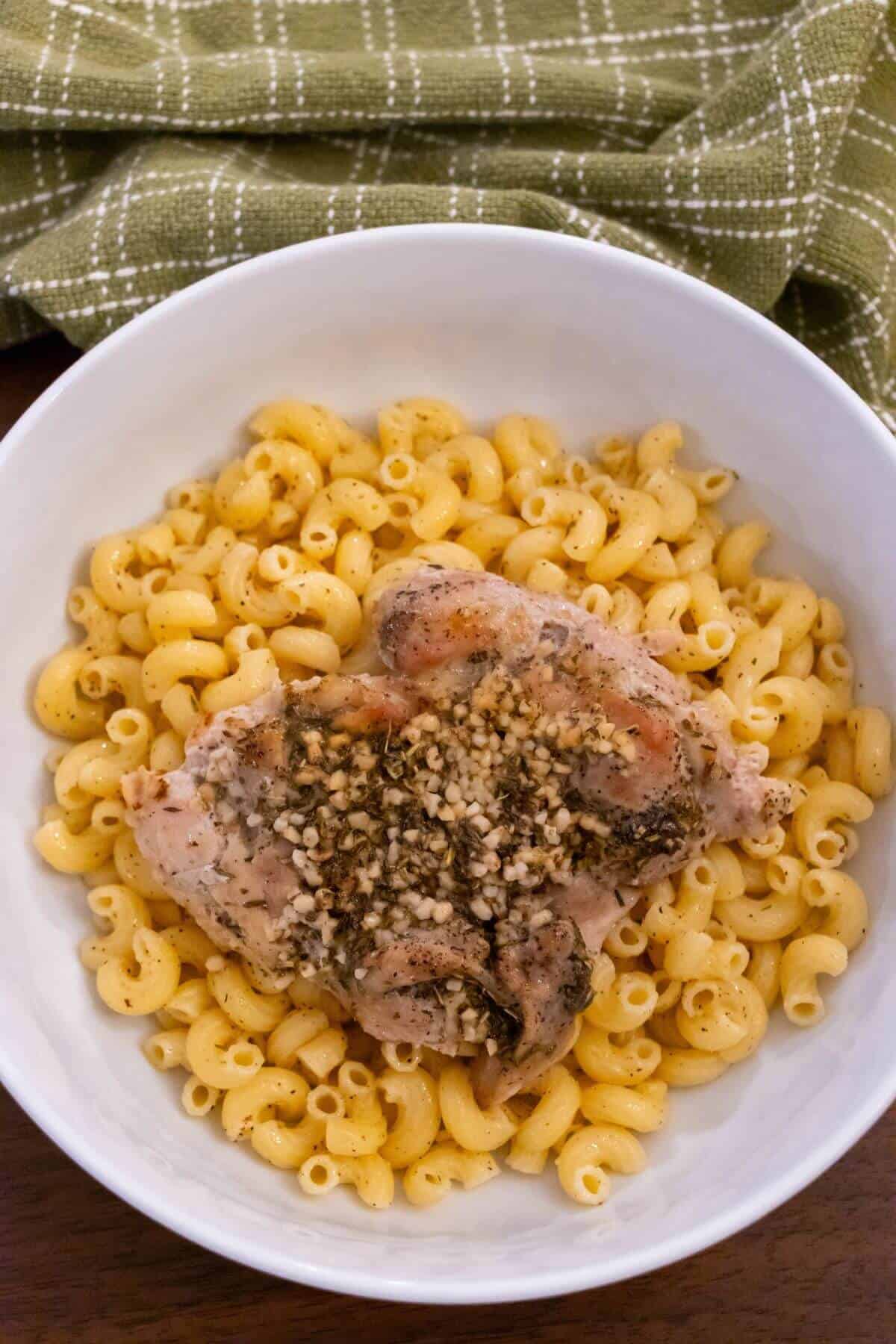 A bowl of macaroni topped with a seasoned, baked chicken thigh, set on a wooden table with a green checkered cloth in the background.
