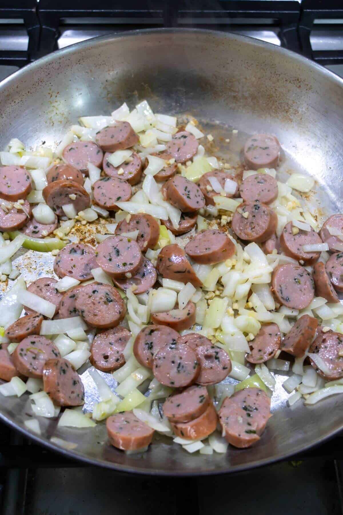 Sliced sausage and chopped onions cooking in a stainless steel skillet on a stovetop.