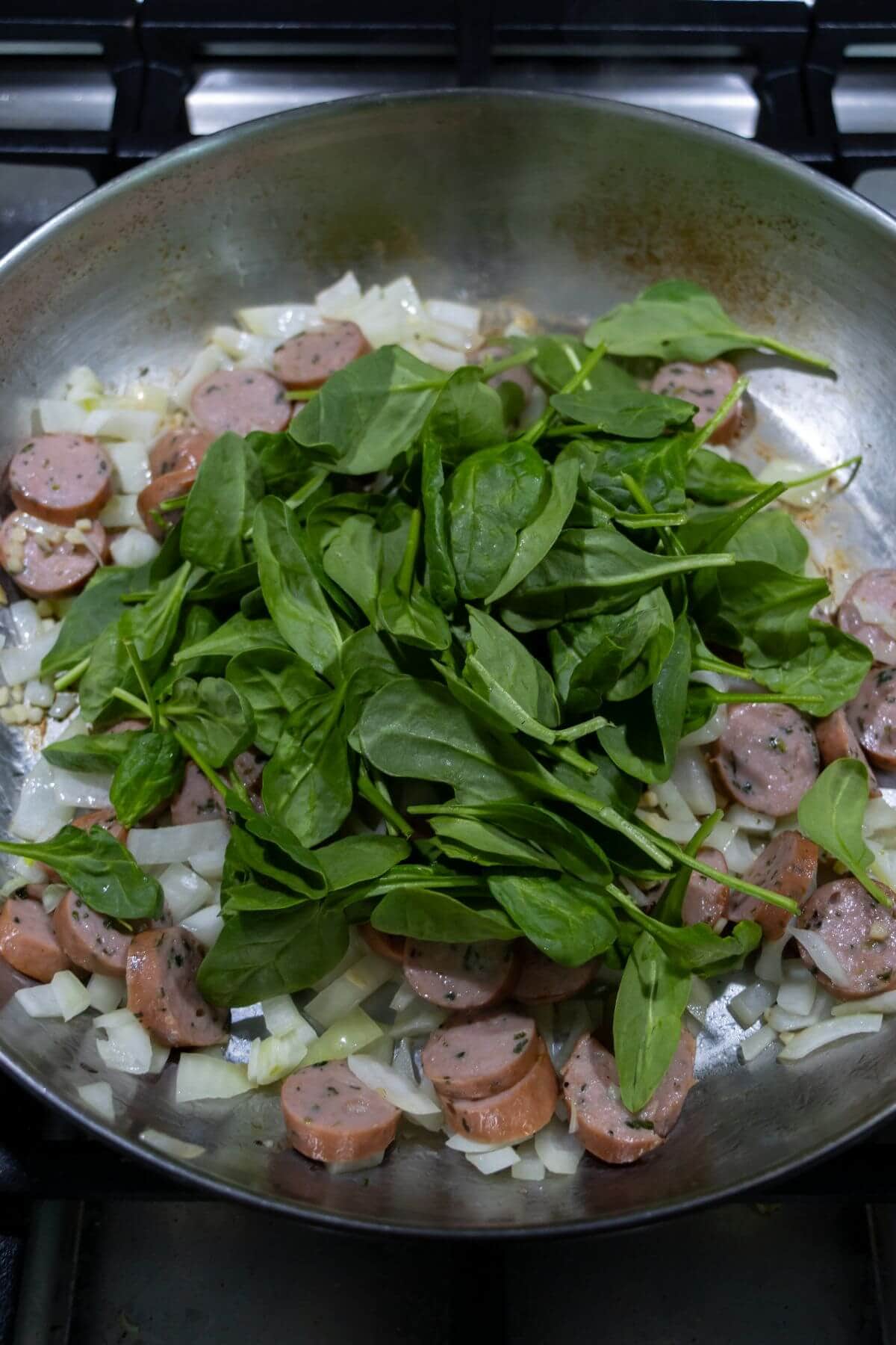 Sliced sausage, chopped onions, and fresh spinach cooking in a stainless steel pan on a stovetop.