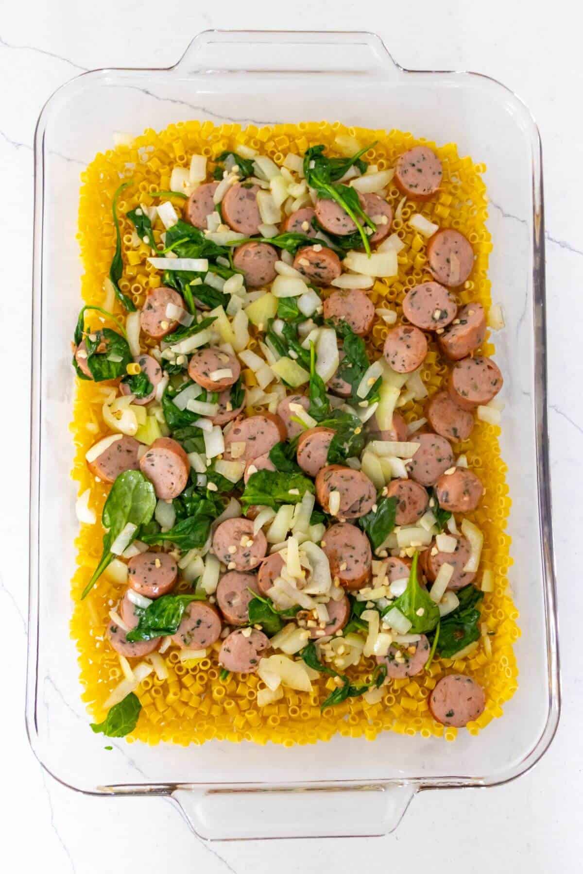 A glass baking dish filled with uncooked pasta, sliced sausage, spinach, and chopped onions on a light countertop.