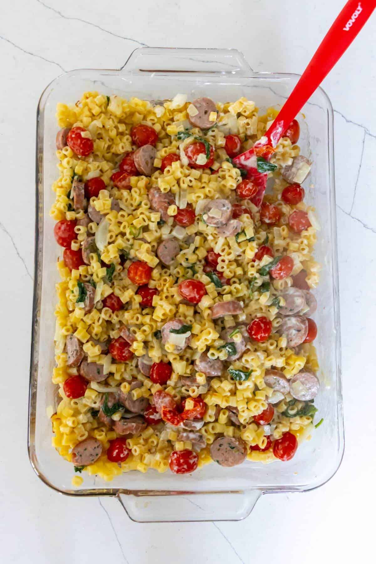 A glass baking dish filled with cooked pasta, sliced sausage, cherry tomatoes, and greens, mixed with a creamy sauce. A red serving spoon is placed in the dish.