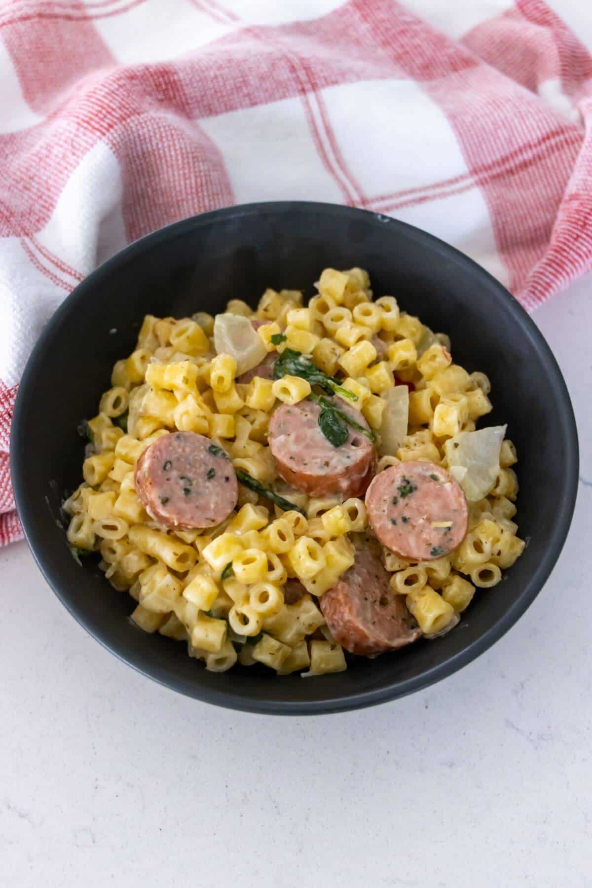 A black bowl filled with macaroni and cheese, sliced sausage, and diced onions, placed on a white surface next to a red and white checkered cloth.