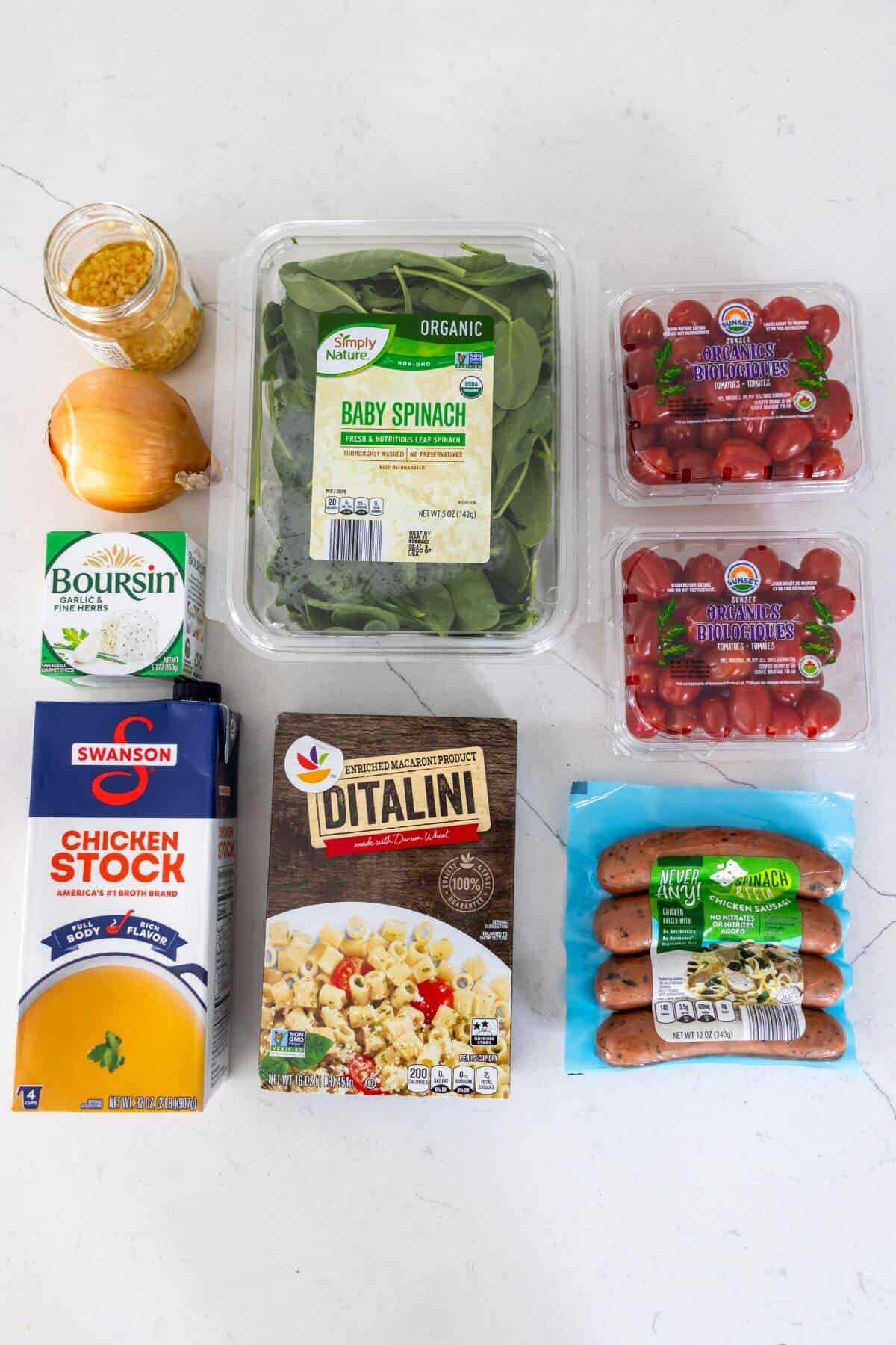 Assorted ingredients on a white surface, including baby spinach, grape tomatoes, chicken stock, ditalini pasta, onion, corn, Boursin cheese, and chicken sausage.