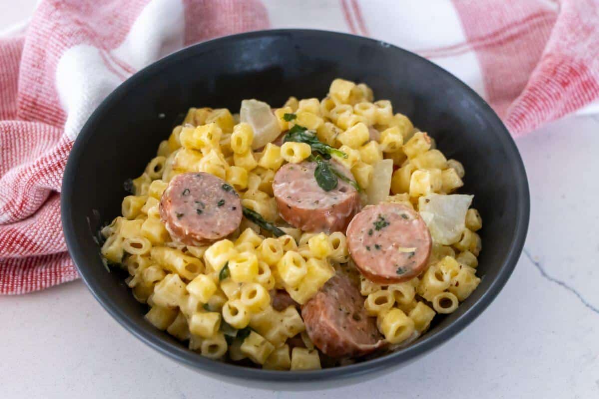A black bowl filled with pasta in a creamy sauce, slices of sausage, herbs, and chopped onions, with a red and white cloth in the background.