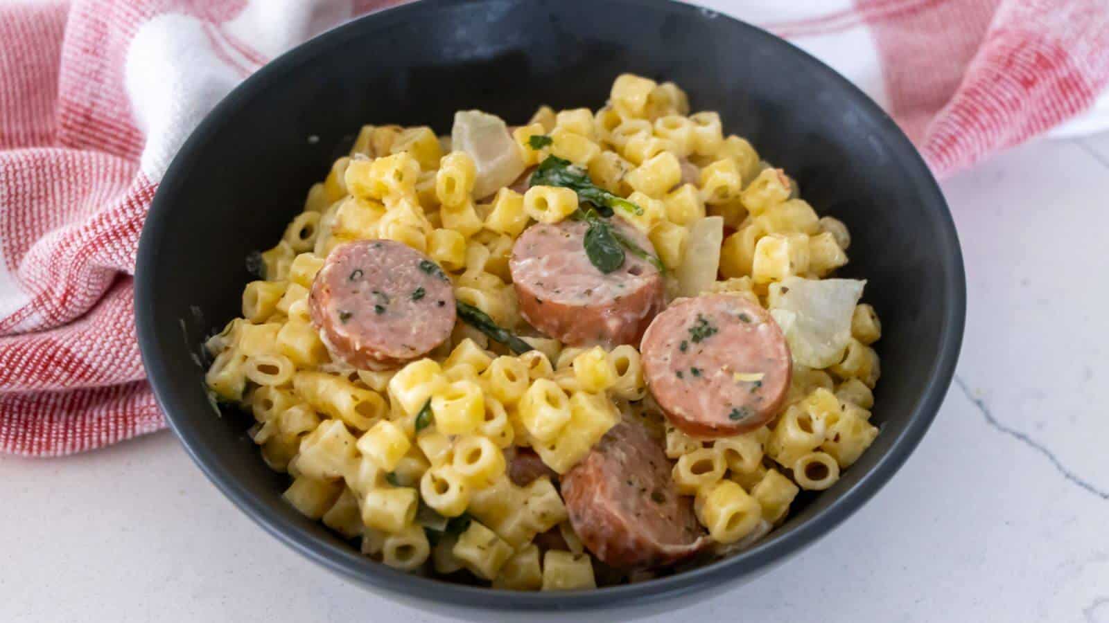 A black bowl filled with creamy pasta and sliced sausage, garnished with herbs and mixed with pieces of onion.