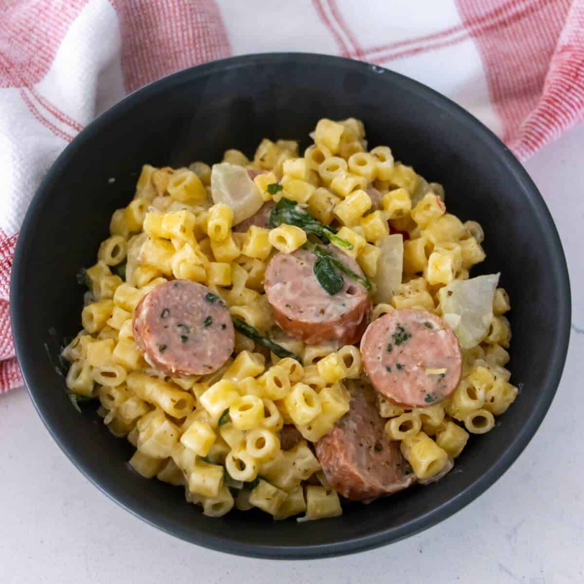 A black bowl filled with macaroni pasta, creamy sauce, sliced sausage, and herbs; red and white towel in the background.