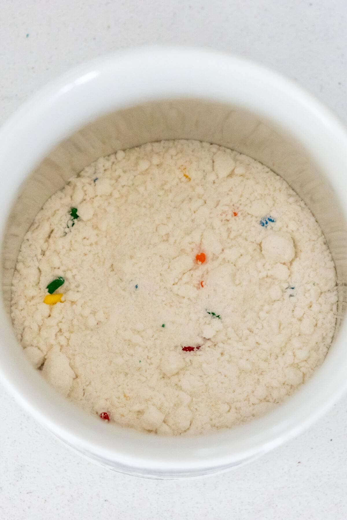 A white bowl filled with off-white cake mix powder, with small colored candy pieces scattered throughout.
