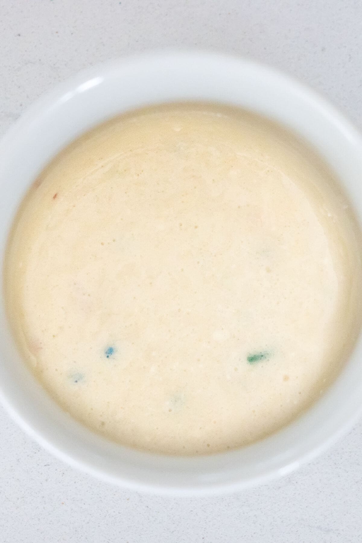 A white bowl filled with a creamy, light-colored sauce with small green and red specks, placed on a light surface.