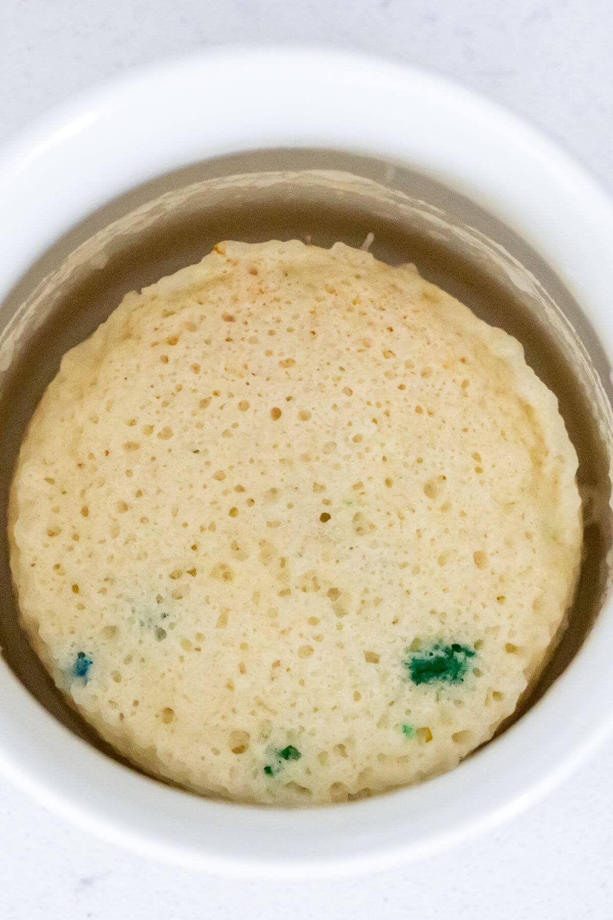 A plain, round mug cake with a few small blue spots, shown inside a white ramekin on a light surface.