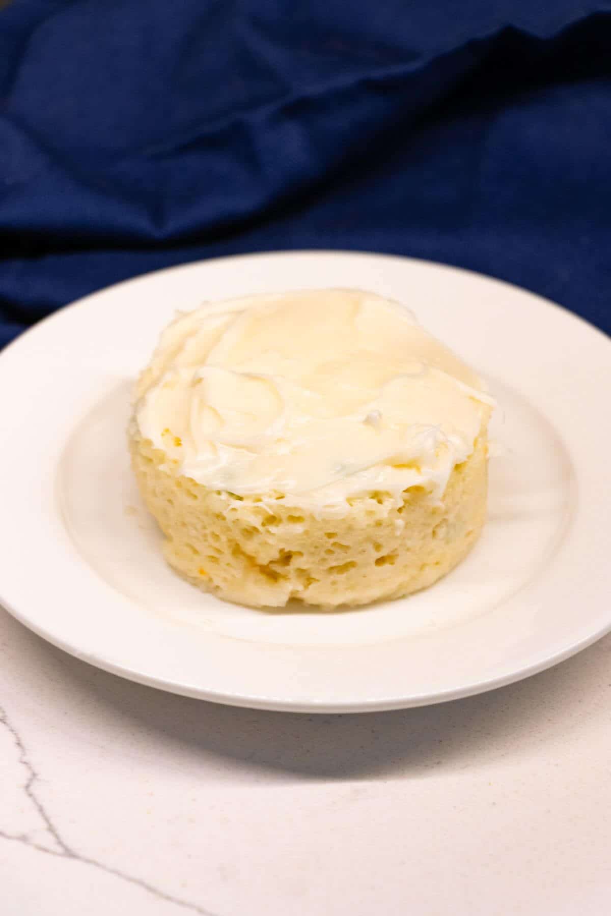 A round, frosted mug cake sits on a white plate against a white surface and a dark blue cloth background.