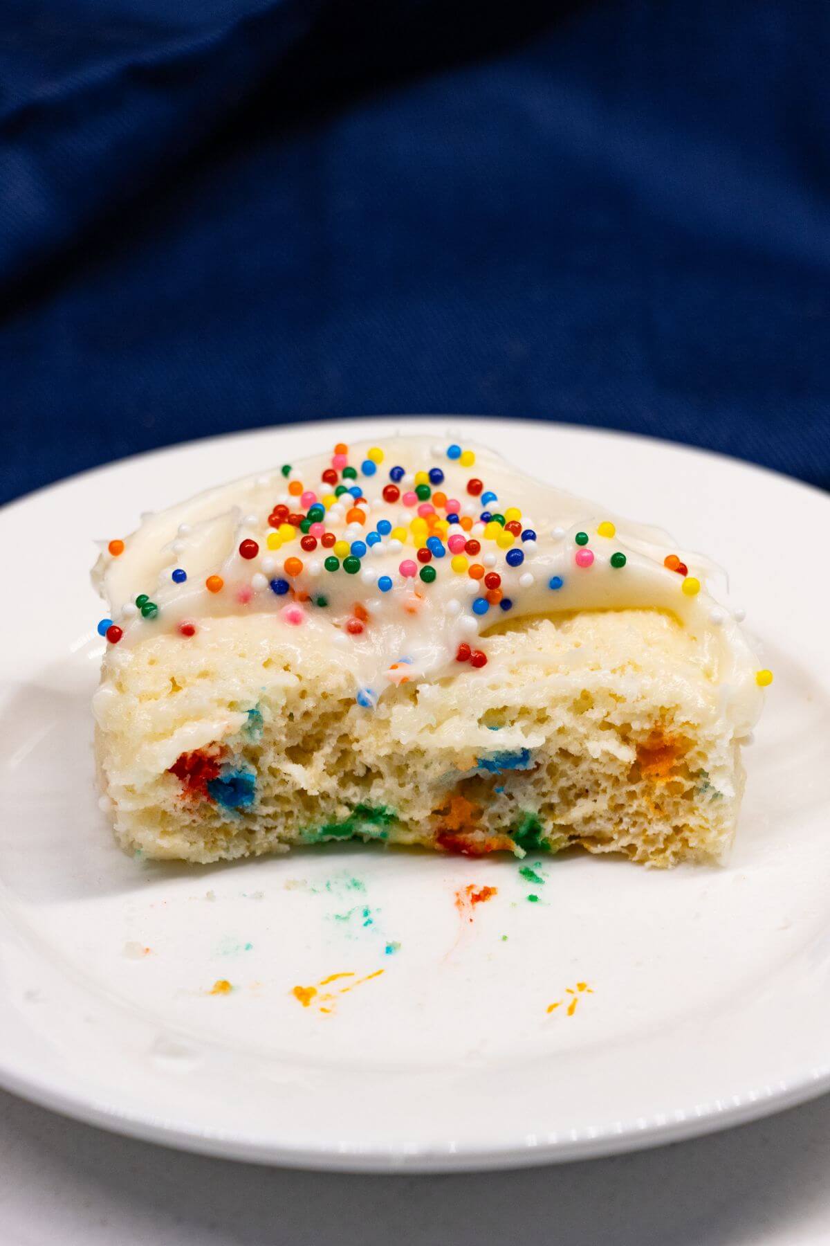A slice of white cake with white frosting and rainbow sprinkles sits on a white plate, with a bite taken out of it.