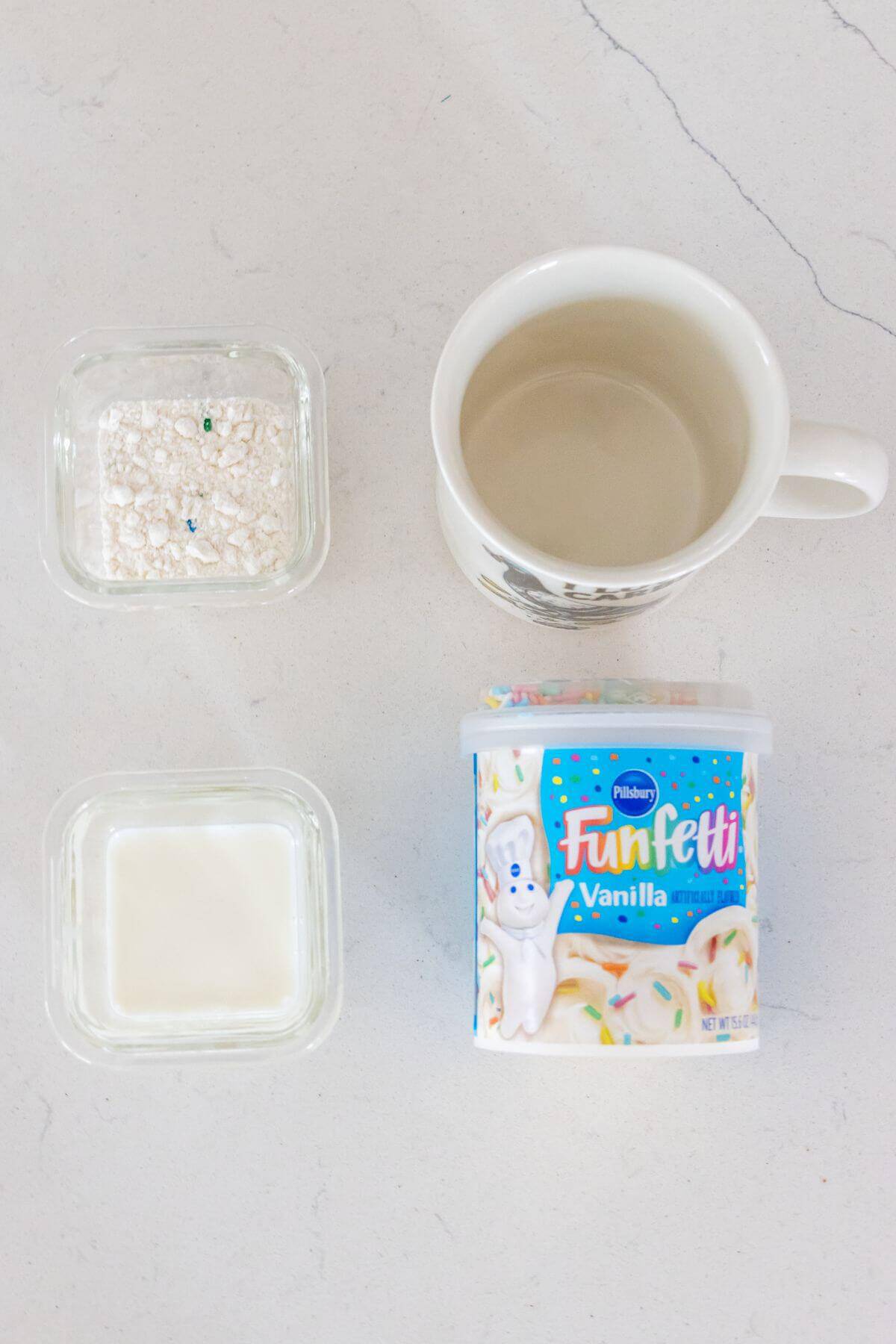 A white mug, glass containers with milk and dry cake mix, a container of Pillsbury Funfetti vanilla frosting, and a cup of water on a white countertop.