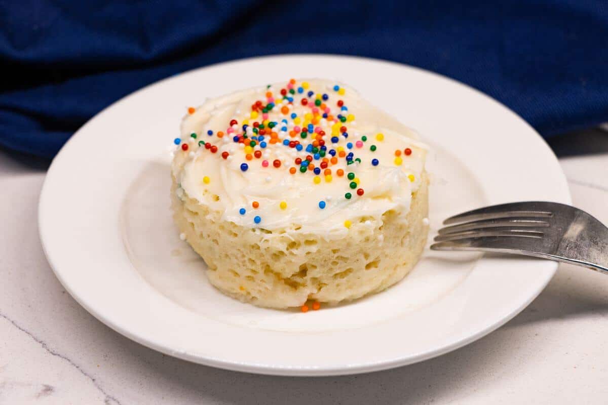 A small vanilla mug cake with white frosting and colorful sprinkles on top, served on a white plate with a fork.