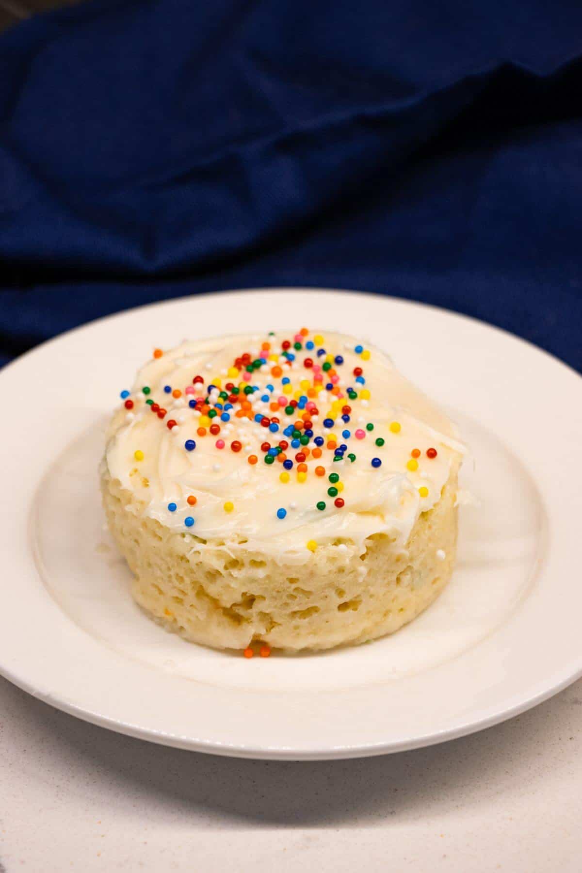 A small vanilla mug cake with white frosting and colorful sprinkles sits on a white plate with a dark blue cloth in the background.