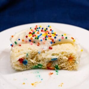 A partially eaten slice of vanilla cake with white frosting and colorful sprinkles sits on a white plate.