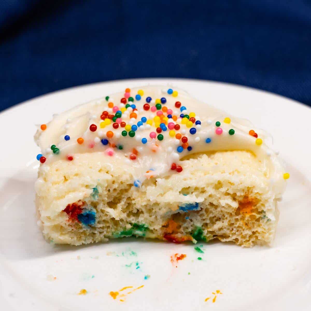 A partially eaten slice of vanilla cake with white frosting and colorful sprinkles sits on a white plate.