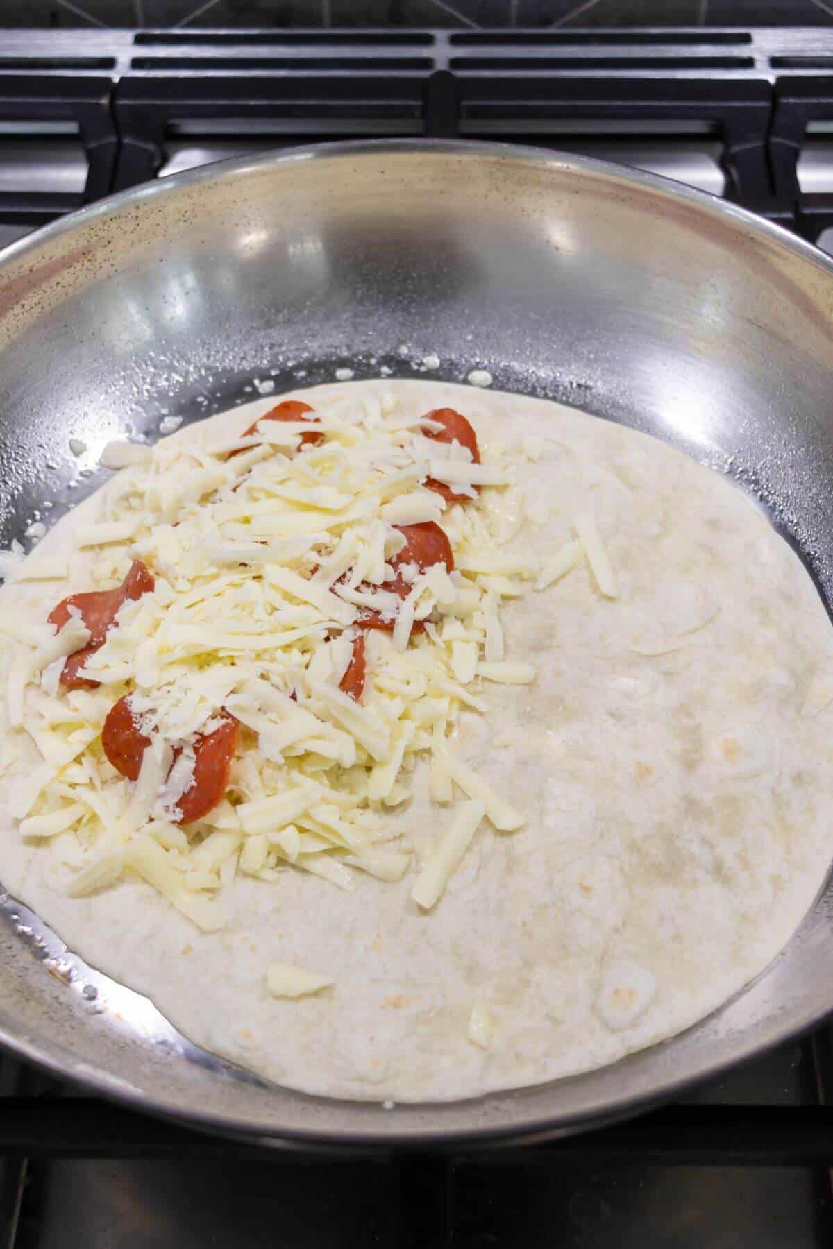 A flour tortilla in a skillet topped with shredded cheese and pepperoni slices is being prepared on a stovetop.