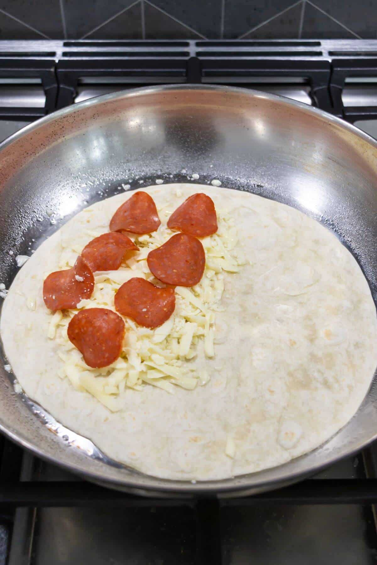 A flour tortilla in a skillet is topped with shredded cheese and pepperoni slices, with one half left plain.