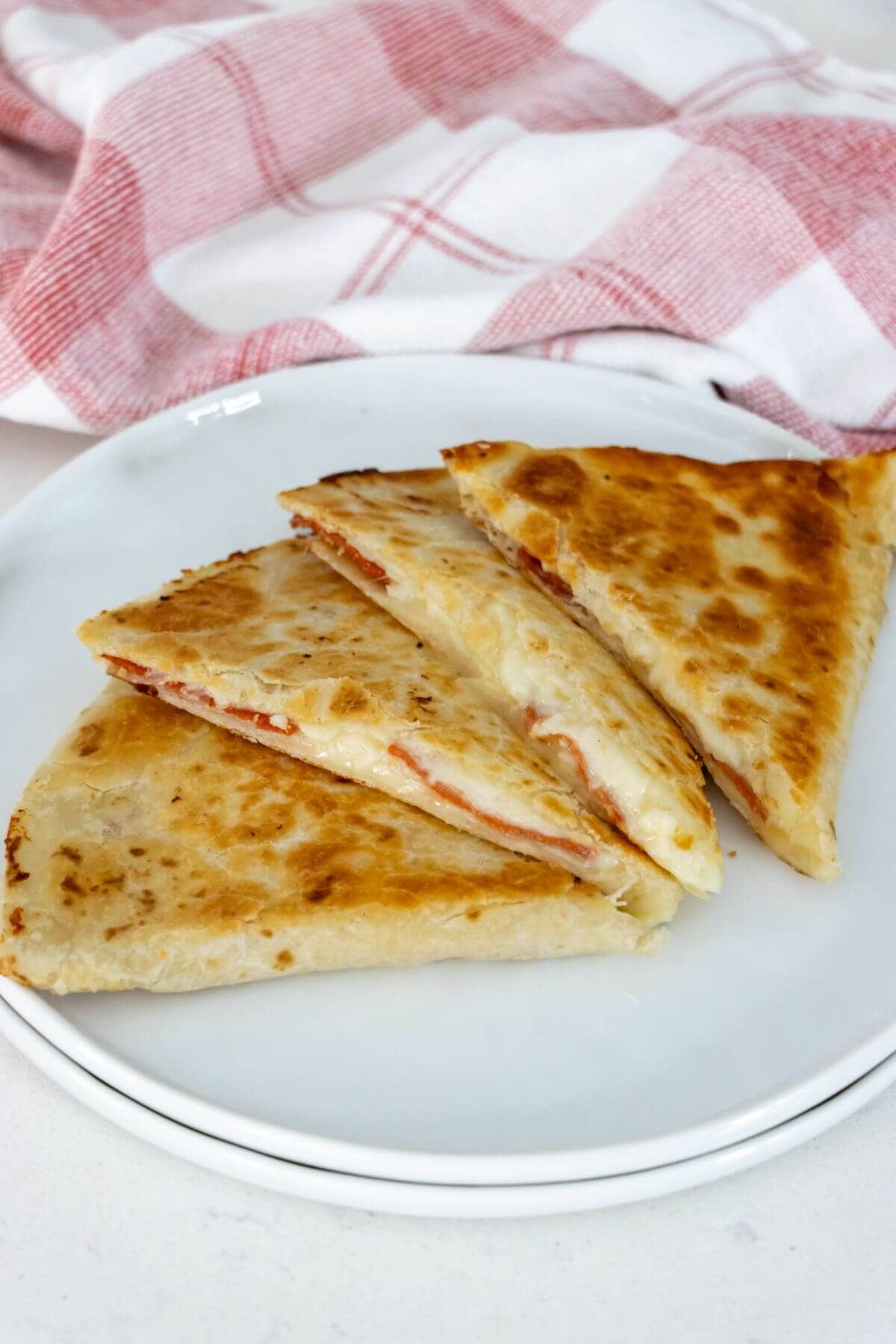 Three triangular slices of a toasted quesadilla with cheese and pepperoni filling are arranged on a white plate, with a red and white checkered cloth in the background.