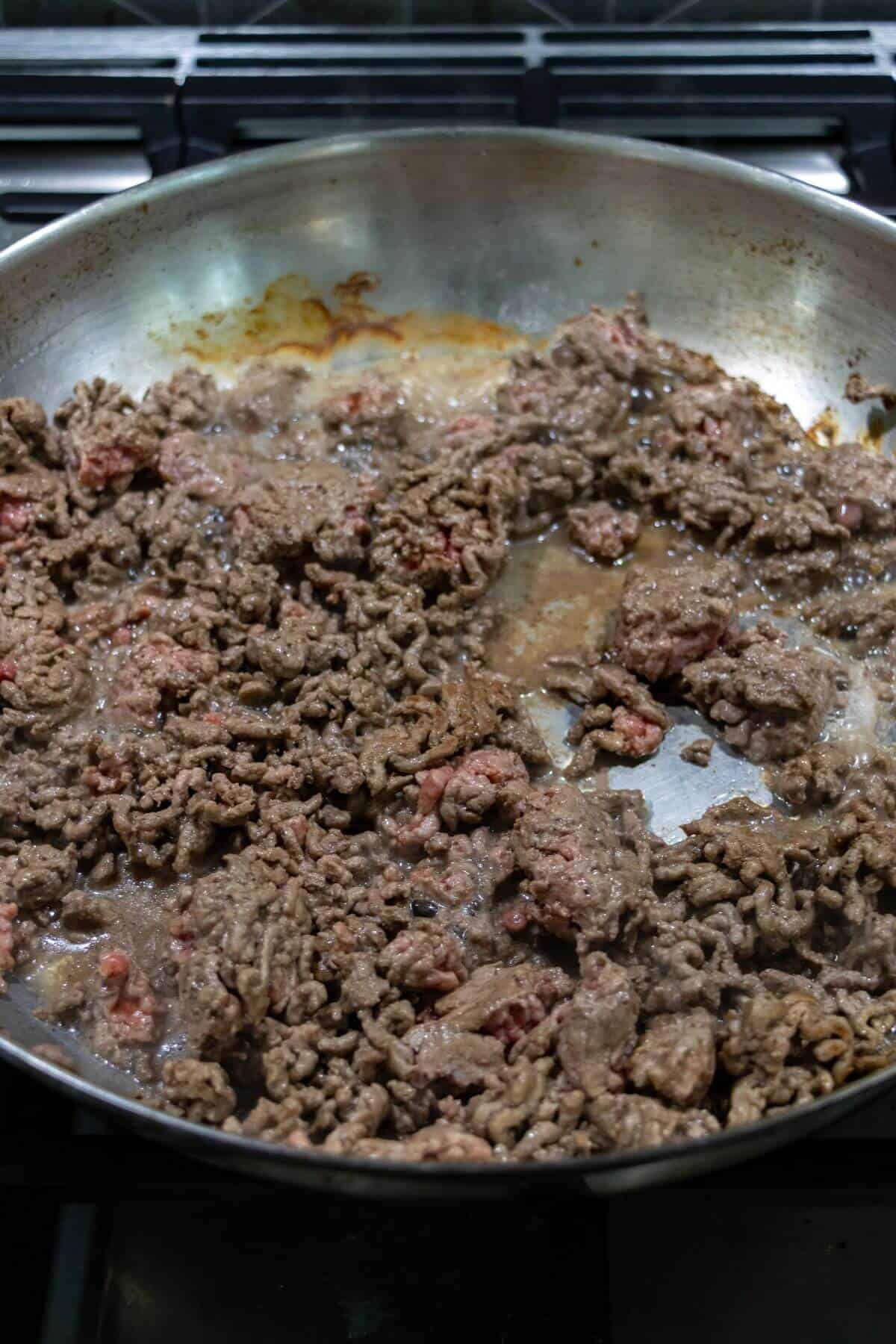 Ground beef cooking in a large metal skillet on a stovetop, with some pieces browned and others still partially pink.