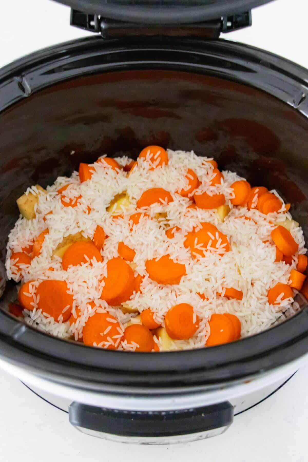 Uncooked white rice, sliced carrots, and pieces of ginger inside an open slow cooker.