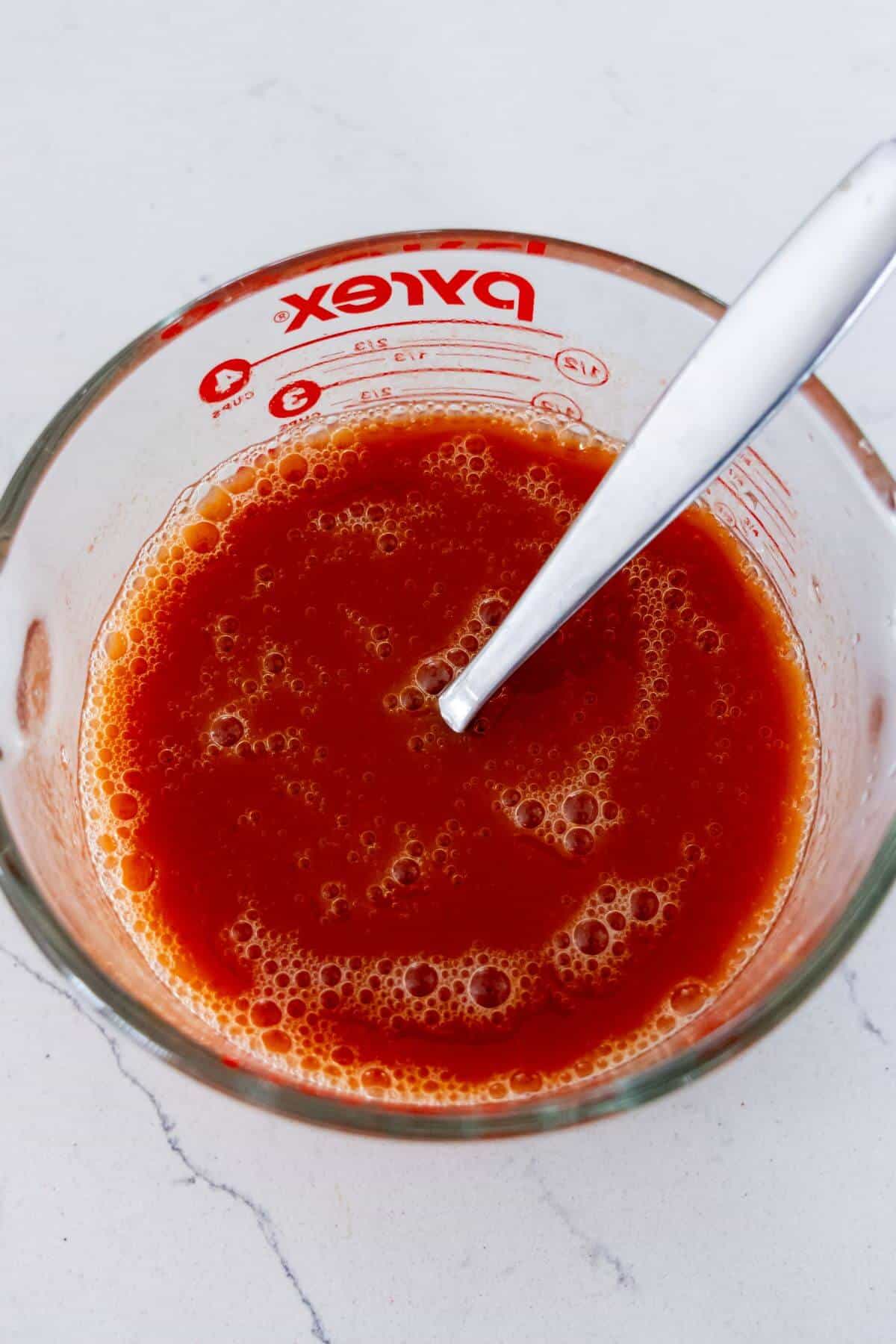 A glass measuring cup filled with red tomato sauce and a metal spoon on a white surface.