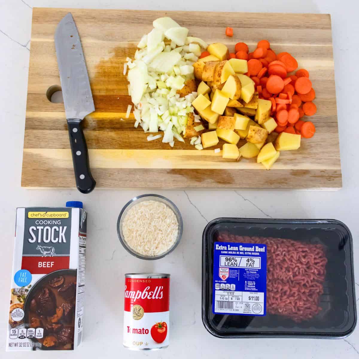 Chopped onions, potatoes, and carrots on a cutting board with a knife, next to beef stock, tomato soup, rice, and extra lean ground beef—everything you need for a hearty slow cooker shipwreck casserole.