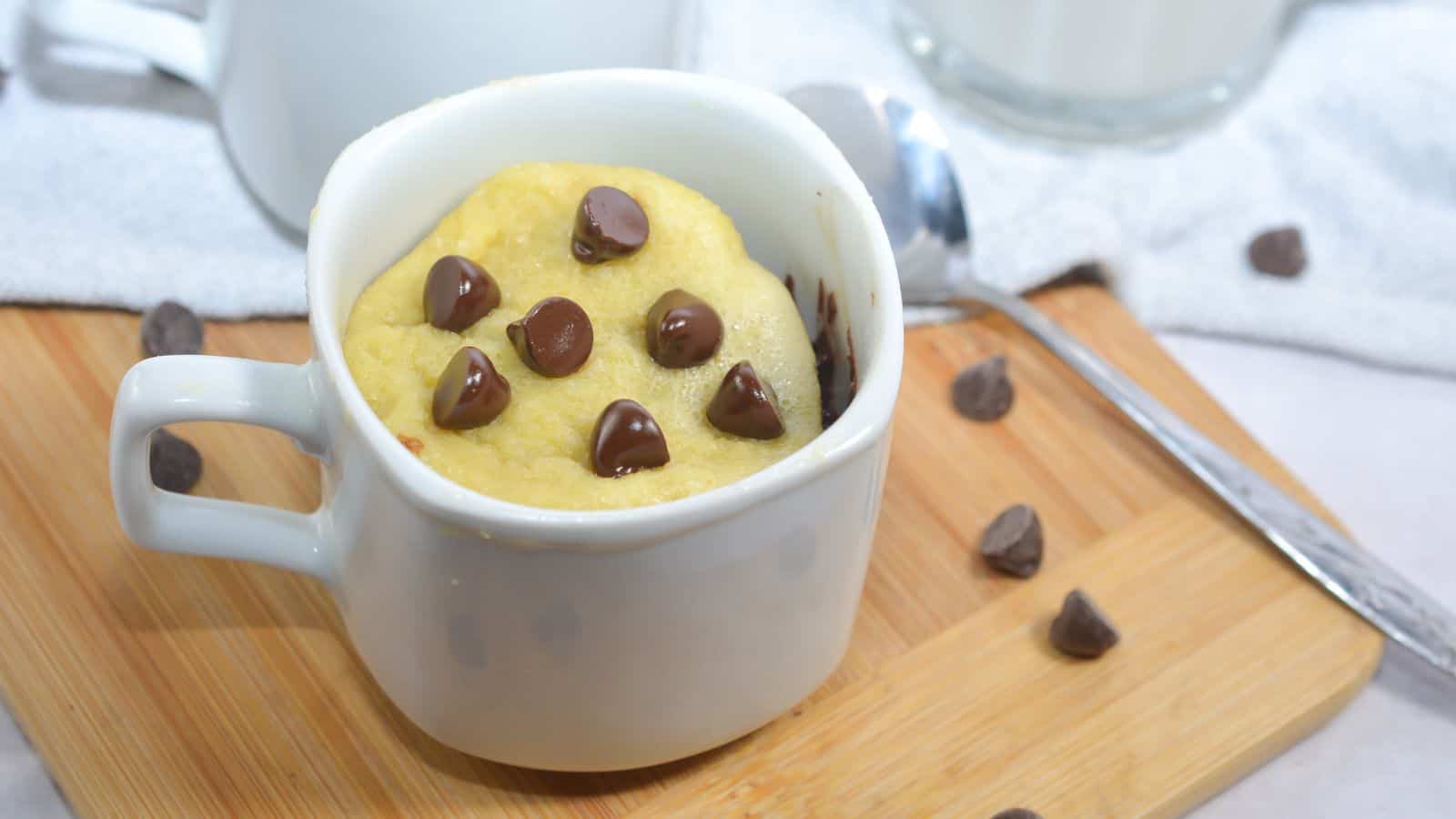 A white mug filled with a baked chocolate chip mug cake sits on a wooden board, with a spoon and scattered chocolate chips nearby.