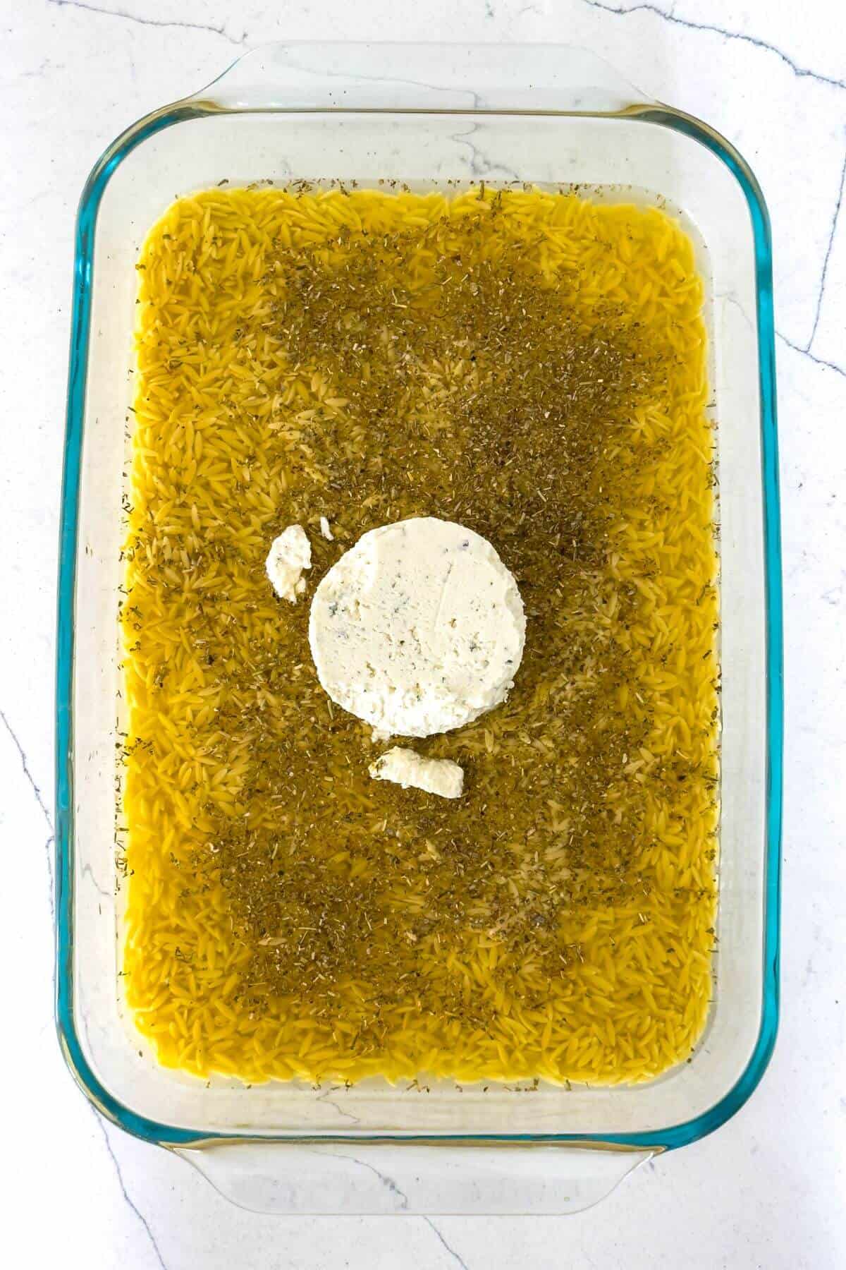 Glass baking dish with uncooked rice, yellow liquid, herbs, and a round block of cheese in the center, set on a white countertop.