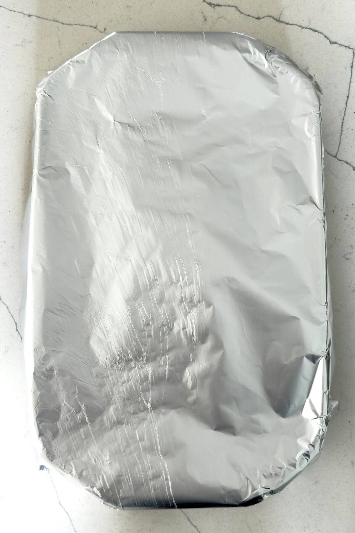 A rectangular baking dish covered tightly with aluminum foil, placed on a light-colored marble countertop.