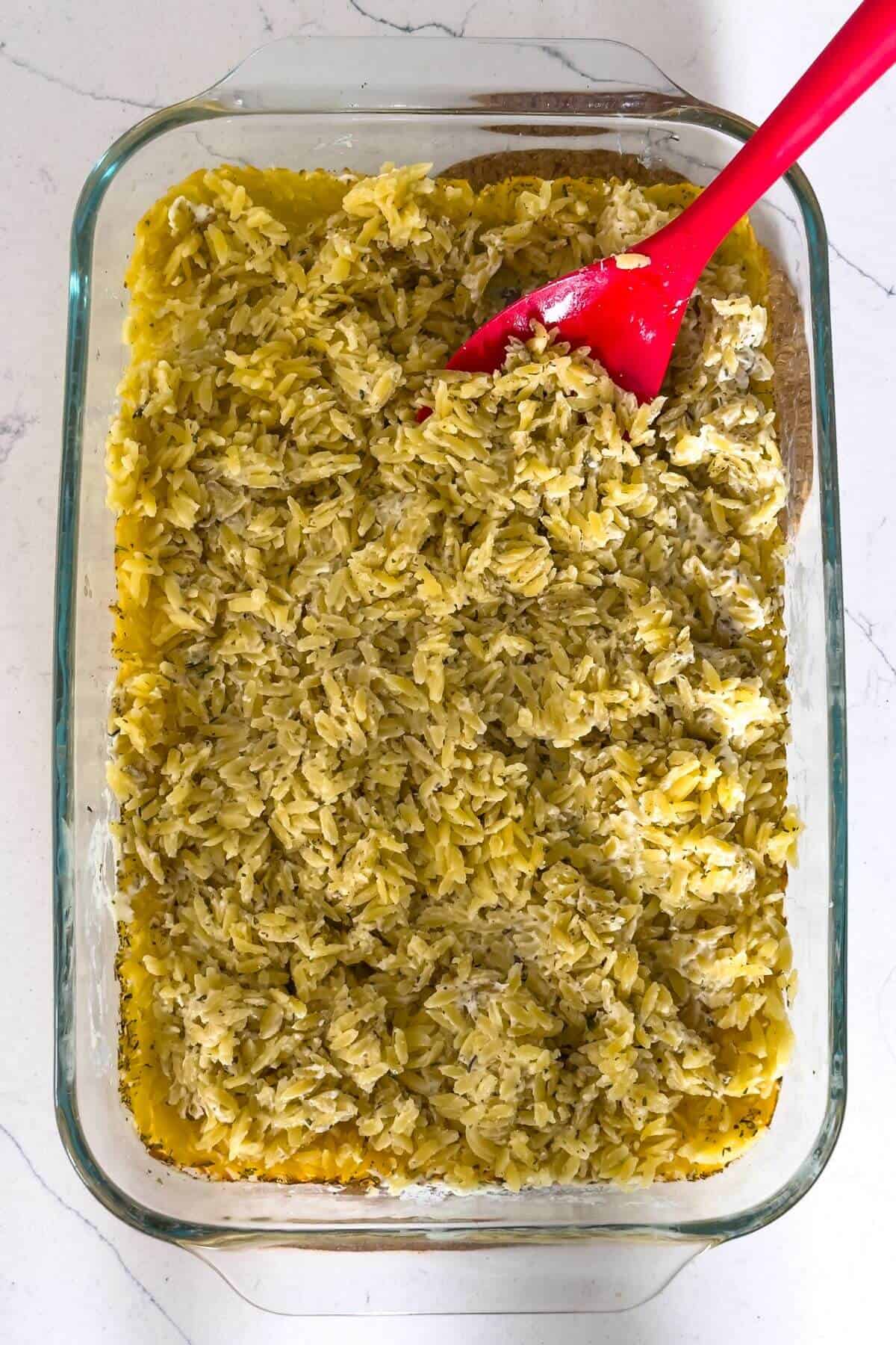 A glass baking dish filled with cooked orzo and rice mixture, being stirred with a red spatula on a white countertop.