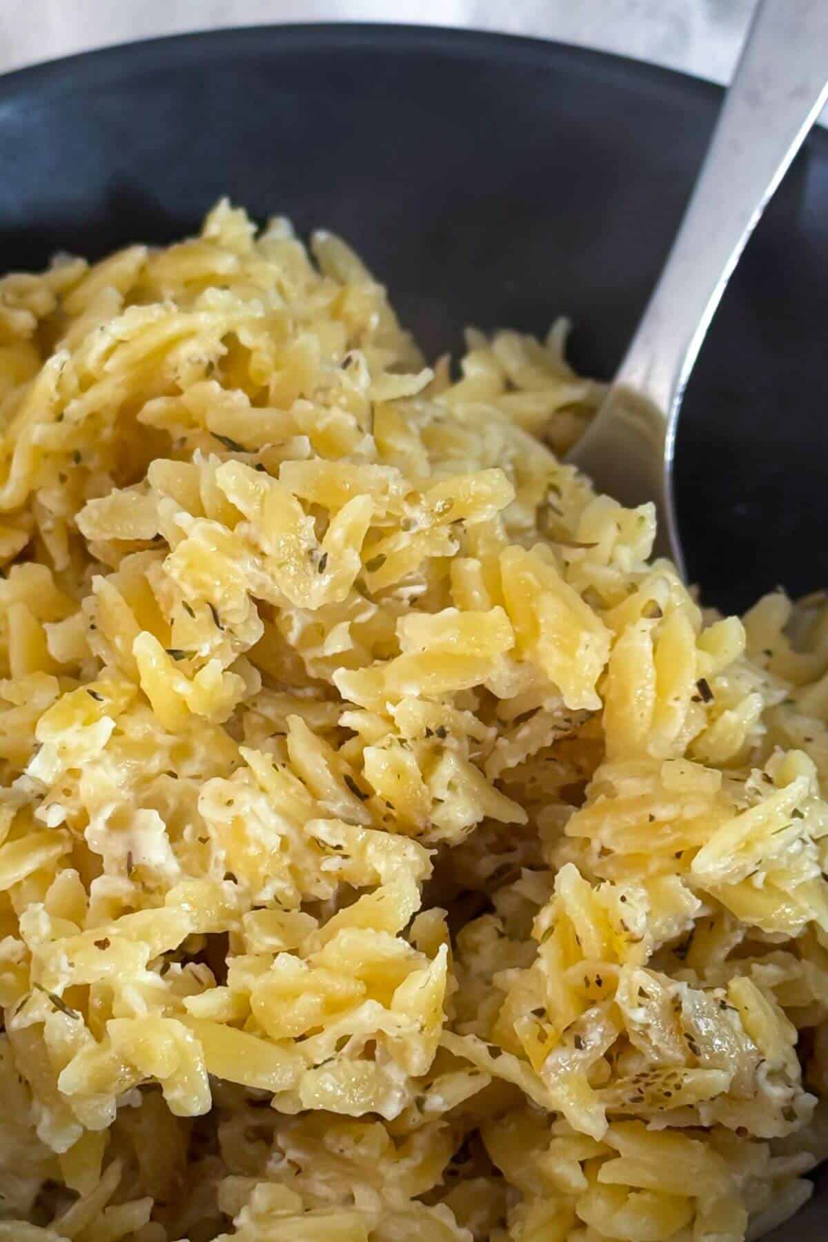 Close-up of cooked orzo pasta mixed with herbs and a creamy sauce, served in a dark bowl with a metal spoon.