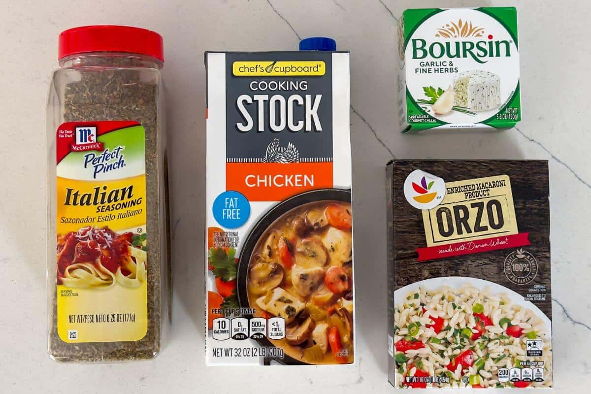 Four food items on a countertop: Italian seasoning, chicken cooking stock, Boursin garlic and herb cheese, and a box of orzo pasta blend.